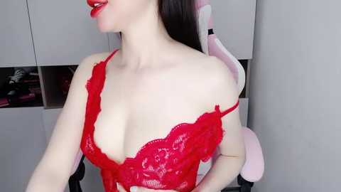 Media: Video of a fair-skinned woman with long dark hair, wearing a red lace bra, sitting in a pink and white gaming chair.