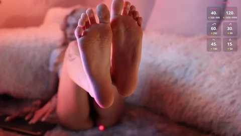 Media: Video of a woman's feet, soles facing the camera, lying on a soft surface, displaying a tattoo on her ankle. Background features a blurred, cozy indoor setting.