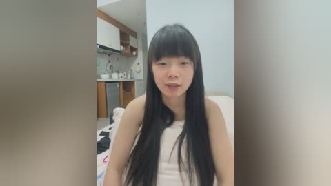 Media: Video of a young Asian woman with long black hair and bangs, wearing a white tank top, sitting in a modern kitchen with wooden cabinets and white countertops.