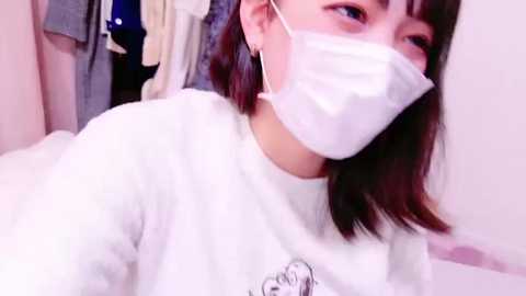 Media: A young Asian woman with shoulder-length brown hair, wearing a white face mask, white sweater, and small hoop earrings, sits indoors in a softly lit room with hanging clothes.