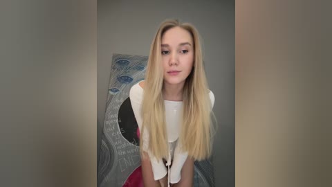 Media: Video of a young woman with long blonde hair, wearing a white blouse, standing in front of a textured, abstract blue wall with circular patterns.