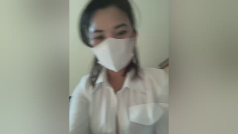 Media: Blurry video of an Asian woman with medium skin tone wearing a white mask, white shirt, and dark hair pulled back, standing in a dimly lit room with beige walls.