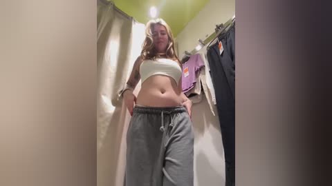 Media: Video of a young woman with light skin and long, blonde hair in a white sports bra and grey sweatpants, standing in a dimly lit, narrow hallway with beige walls and a purple towel hanging on a rack.