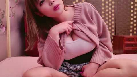 Media: Video of a young woman with fair skin and long brown hair, wearing a pink sweater over a white tank top, sitting on a bed.