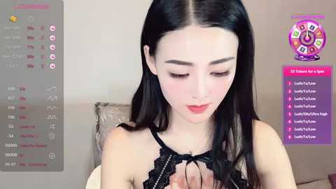 Media: Video of a fair-skinned, slender Asian woman with long black hair, wearing a black lace halter top, sitting on a beige couch. Background shows a digital clock and a colorful spinning wheel game.