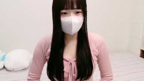 Media: Video of a young Asian woman with long black hair and bangs, wearing a white face mask and a pink top. She is kneeling on a striped bed with white pillows in a simple, white room.