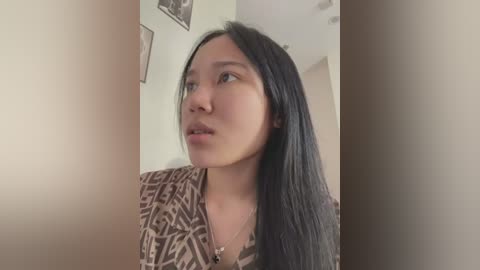 Media: A video of an East Asian woman with long black hair, fair skin, and brown patterned top, looking upward with a thoughtful expression in a dimly lit room with beige walls and framed pictures.