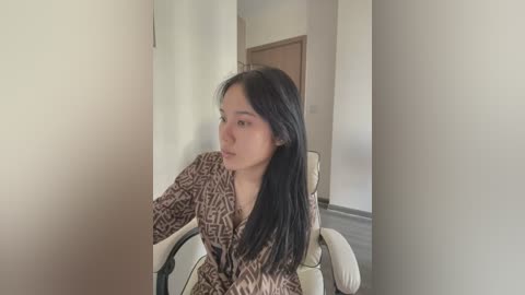 Media: Video of an Asian woman with long black hair, wearing a leopard print robe, sitting in a beige armchair in a minimalist room with white walls and wooden floor.