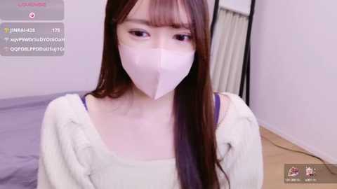 Media: Video of an East Asian woman with long, straight brown hair, wearing a white mask and a cozy off-white sweater, standing in a simple bedroom with light-colored walls.