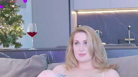 Media: Video of a fair-skinned, blonde woman with wavy hair, wearing a white top, sitting on a beige couch in a modern kitchen. Background features a Christmas tree, wine glass, and sleek, minimalist decor.