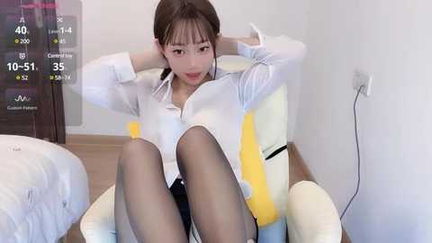 Media: Video of an Asian woman with light skin, wearing a white blouse and black skirt, sitting on a yellow armchair, legs crossed, in a minimalist, white-walled room.