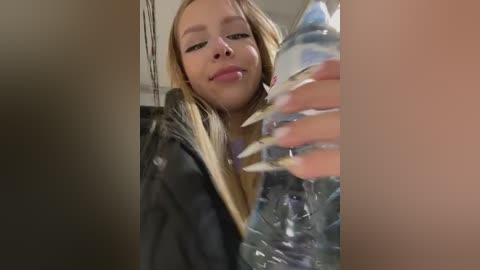 Video of a young Caucasian woman with light skin and long blonde hair, wearing a black jacket, pouring water from a plastic bottle.