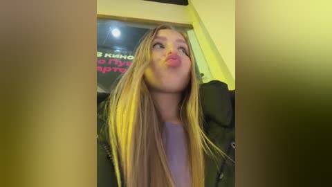 A video of a blonde woman with straight hair, wearing a purple top, puckering her lips in a playful manner, taken indoors with a yellowish-green filter.