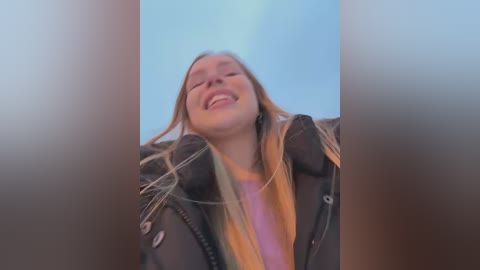 Media: Video of a smiling young woman with long blonde hair, wearing a black jacket and pink shirt, against a clear blue sky, taken from a low angle.