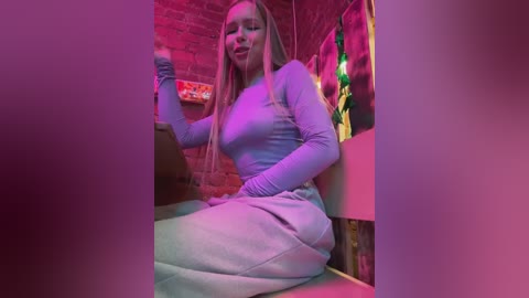 Video of a blonde woman with long hair, seated on a couch, wearing a tight, light purple sweater and pants, in a dimly lit room with red and purple lighting, holding a drink.