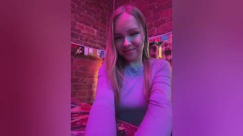 Video of a young blonde woman with fair skin, wearing a light blue shirt, grinning with her hands clasped, sitting on a bed in a cozy room with a brick wall and pinkish-purple lighting.