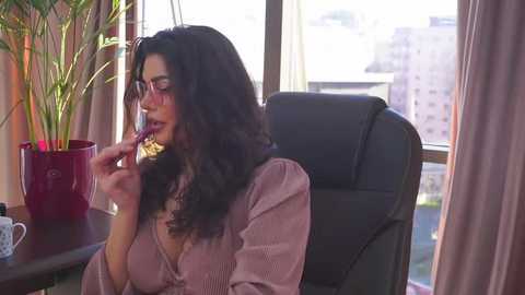 Media: Video of a woman with long, dark hair, wearing glasses, and a pink blouse, eating a lollipop, sitting in a black office chair by a window with a city view.