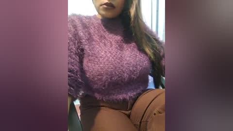 Media: Video of a woman with medium brown skin and long brown hair, wearing a fuzzy, purple turtleneck sweater and brown, high-waisted pants, seated indoors with blurred background.