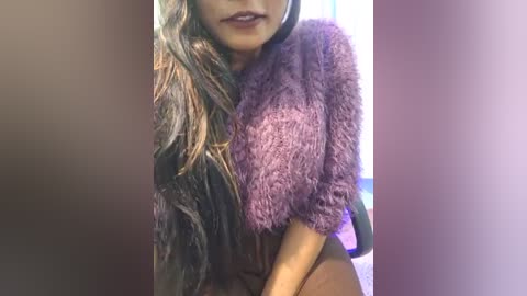 Media: Video of a woman with long, wavy brown hair, wearing a fuzzy, purple, short-sleeved sweater. Her lips are painted a deep burgundy. Background features a blurred, indistinct room with a hint of purple lighting.