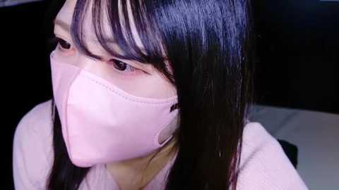 Media: Video of a young Asian woman with long black hair and bangs, wearing a pink face mask, white shirt, and dark surroundings.