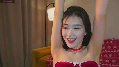 Media: Video of an East Asian woman with long black hair, fair skin, and red lingerie, stretching her arms above her head in a bedroom with a red-patterned blanket and a lamp.