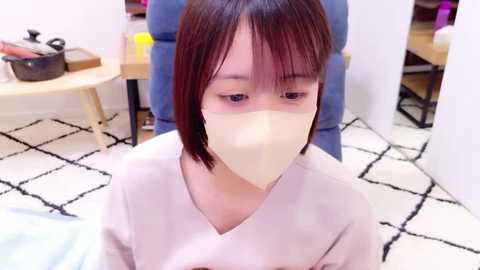 Media: Video of a young East Asian woman with straight, shoulder-length brown hair, wearing a white surgical mask and light pink scrubs, seated in a blue upholstered chair. Background shows a modern office with a black and white geometric-patterned rug, wooden table, and purple objects.