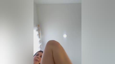 Media: Video of a nude woman with light skin, dark hair, and a slender physique, lying on her back with her legs raised. The background features a minimalist, white-walled room with a modern, white wall-mounted lamp.