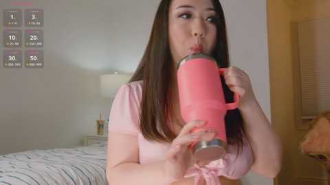 Media: A video of an Asian woman with straight black hair, wearing a pink top, drinking from a pink thermos, in a modern bedroom with a white bed and nightstand.