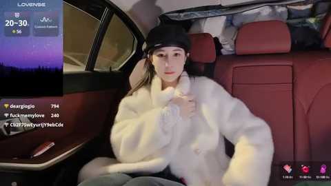 Media: A video of a woman in a plush white fur coat and black beanie, seated in a red car, with a cityscape view in the background.