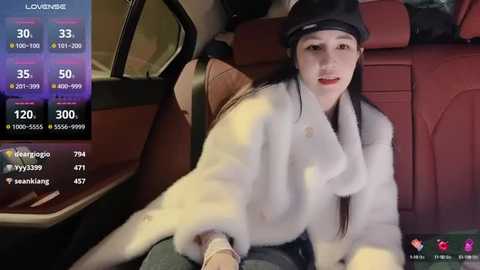 Media: Video of a young Asian woman with pale skin, long black hair, and light makeup, wearing a white fur coat and black hat, seated in a red leather car seat.