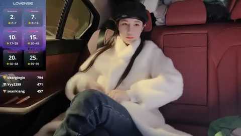 Media: Video of a young Asian woman in a car, wearing a black headband, white fur coat, and jeans, with a digital interface showing stats and names.