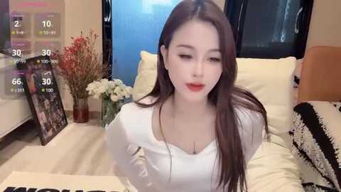 Media: A video of an East Asian woman with long, straight brown hair, wearing a white long-sleeve top, sitting on a beige bed. The background features a floral arrangement, a framed photo, and a black-and-white pillow.