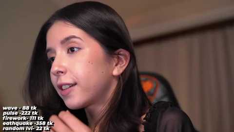 Media: Video of a young woman with long dark hair, light skin, and small mole on her cheek, smiling slightly, in a cozy indoor setting.