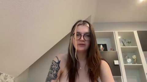 Media: Video of a young woman with long brown hair, glasses, and tattoos, standing in a modern, minimalist room with white shelves and a beige ceiling.