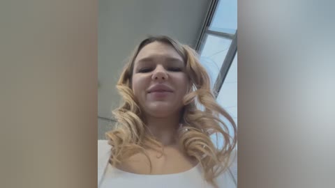 Media: Video of a blonde woman with wavy hair, wearing a white top, standing in an indoor setting with light-colored walls and a window. Her expression is neutral, and the image is taken from a low angle.