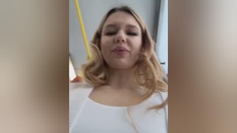 Media: A video shows a young woman with long, wavy blonde hair, fair skin, and closed eyes, wearing a white top. She stands in a bus, with a blurred background and yellow seat.