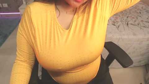 Media: Video of a woman with fair skin and dark hair, wearing a tight, mustard-yellow long-sleeved top that accentuates her large breasts. She sits on a black office chair, in a room with a bed covered in a floral duvet.