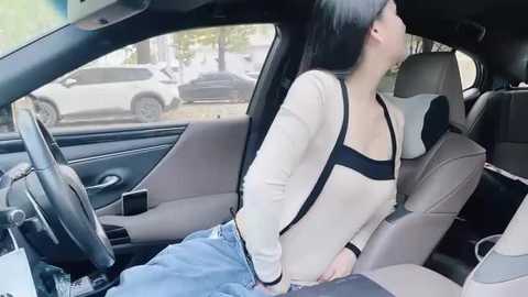 Media: Video of a young woman with long black hair, wearing a beige cardigan and blue jeans, sleeping in a car seat, with a parked car visible outside.