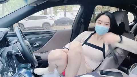 Media: Video of a young East Asian woman in a car, wearing a blue mask, beige top, and shorts, with a gray backpack, sitting in a gray seat, holding a smartphone.