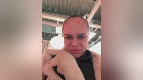 Media: Video of a bald, light-skinned man with a fur coat, wearing a dark shirt, frowning in a warehouse with exposed beams and white lights.