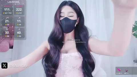 Media: A video of an Asian woman with long, wavy black hair, wearing a black face mask, a light pink lace top, and a silver necklace, in a modern, white room.