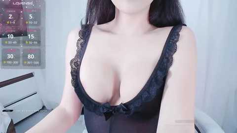 Media: Video of a fair-skinned woman with long black hair, wearing a revealing black lace lingerie top that highlights her ample cleavage. Background features a calendar and white walls.