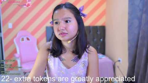 Media: Video of an Asian woman with shoulder-length black hair adorned with pink bows, wearing a purple camisole, seated indoors against a striped wall, with text overlay, \"22 extra tokens are greatly appreciated.\