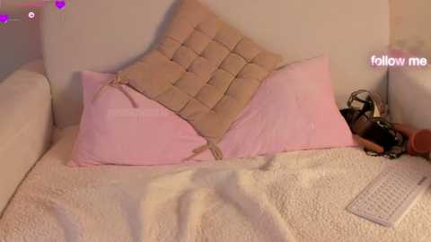 Media: Video of a cozy, pastel-themed bedroom with a white bed, pink pillows, beige tufted cushion, and a plush blanket. A white keyboard rests on the bed, adding a modern touch.