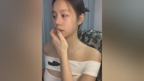 Media: Video of a young Asian woman with fair skin, dark hair in a bun, wearing a white off-shoulder top with a black mustache pattern. She's indoors, with a blurred background showing a patterned chair.