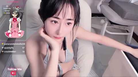 Media: A video of a young East Asian woman with fair skin and long black hair, wearing a grey bikini, sitting on a white chair, with a headset on, in a modern room.