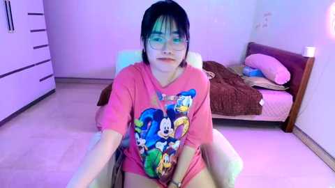 Media: Video of a young Asian woman with short black hair, glasses, and a pink Mickey Mouse T-shirt, sitting on a bed in a minimalistic bedroom with white walls and wooden furniture.