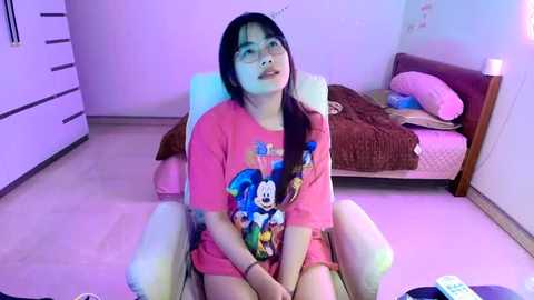 Media: Video of an Asian woman with long black hair, glasses, wearing a pink Mickey Mouse t-shirt, sitting on a white chair in a purple-lit bedroom with a bed and dresser.