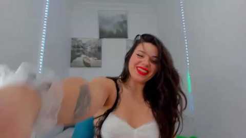 Media: Video of a smiling, red-lipstick-wearing woman with long dark hair, wearing a white lace bralette, standing in a brightly-lit room with white walls, a framed black and white photo, and a bed in the background.