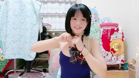 Media: Video of an Asian woman with short black hair, wearing a blue tank top, adjusting a plaid bow tie in a cluttered sewing room with floral fabrics, a chair, and a pink storage bin.